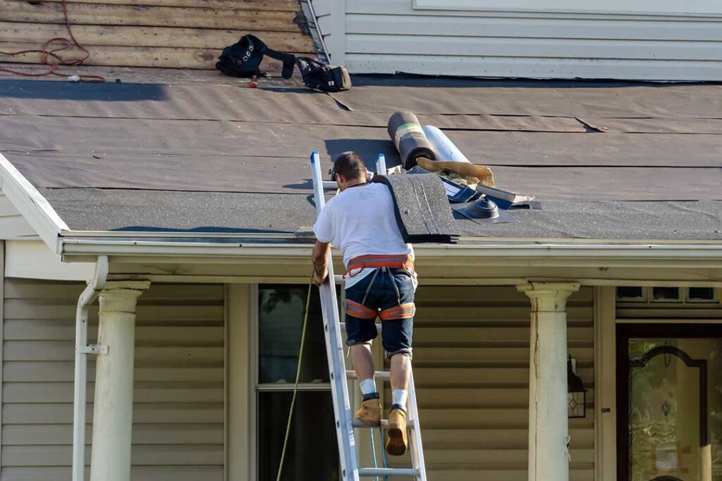 Roofing worker rolls underlayment across a wooden roof deck beneath rows of shingles.