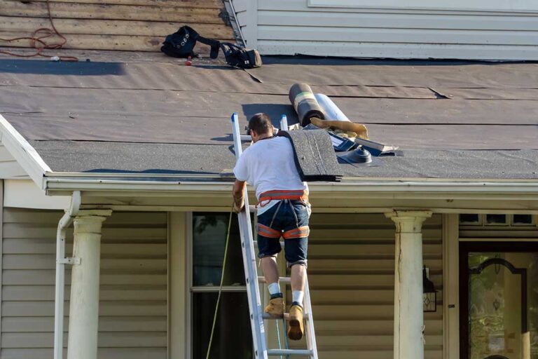 Roofing worker rolls underlayment across a wooden roof deck beneath rows of shingles.