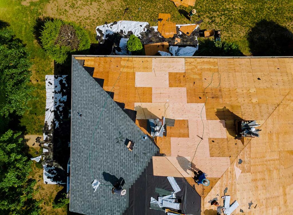 A homeowner examines aging asphalt shingles while inspecting a roof near a leafy residential home.